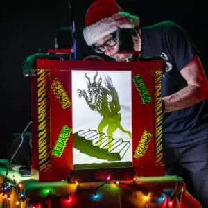 A man in a Santa hat manipulates a shadow puppet of a monster.