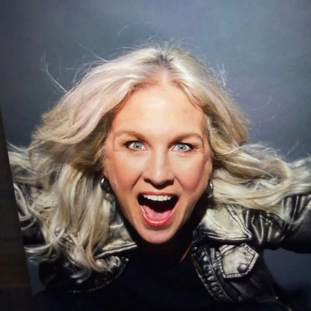 A white woman with silver hair in a black leather jacket stares out at the viewer with her mouth open.