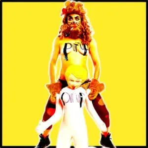 A topless person in red underwear, wearing a lion-style wig, beard, and paw gloves, stands against a yellow background holding an inflatable doll. The words ‘Put ’em Up’ are written on both their chests.