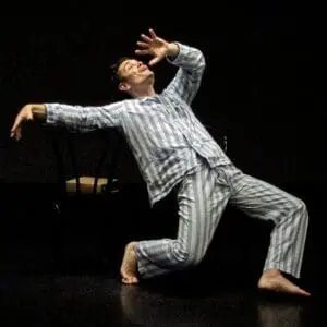 A solo performer in striped pajamas kneels dramatically on a dark black-box stage, reaching upward beside two shadowed chairs in a gothic scene inspired by Edgar Allan Poe’s Ligeia.