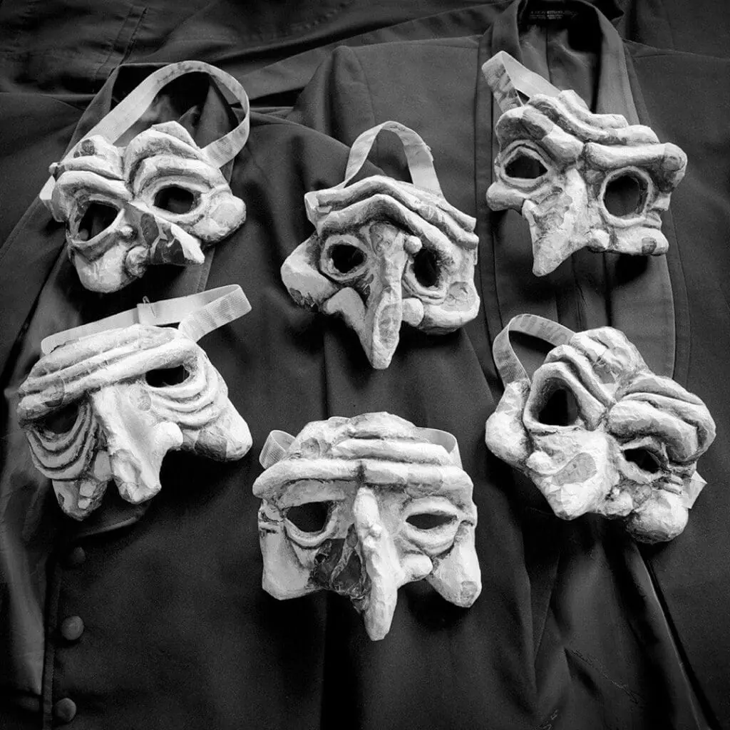 Six Surrealist masks with furrowed brows, dark hollow eyes, and large noses set against black dress coats.