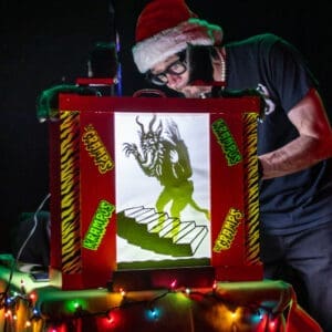 A man in a Santa hat manipulates a shadow puppet of a monster.